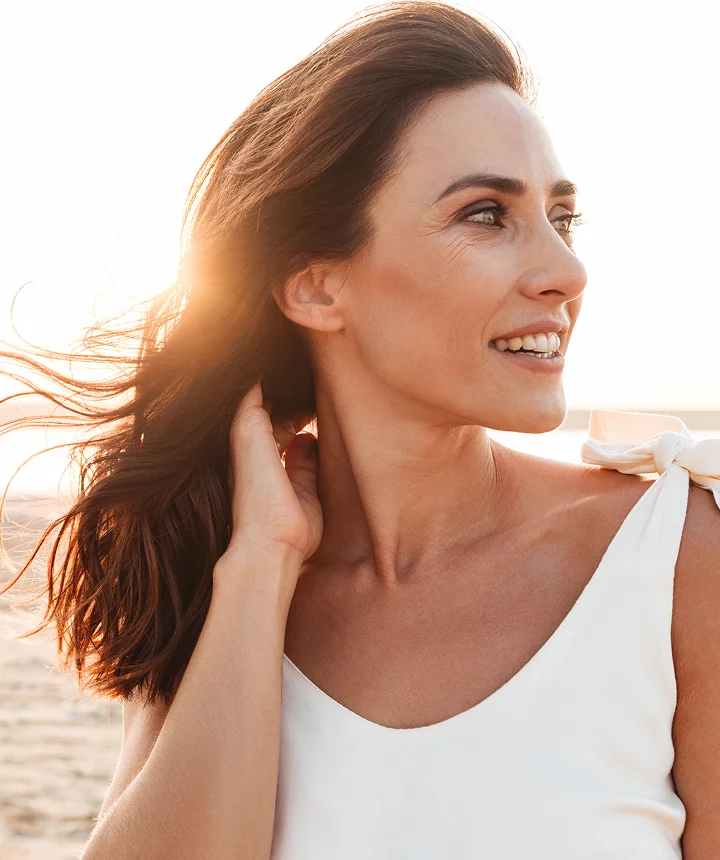 A bright, sun-drenched portrait of a smiling woman with brunette hair looking off to the side, wearing a white sleeveless top with a bow detail on the shoulder. - Lasers in Los Angeles, CA