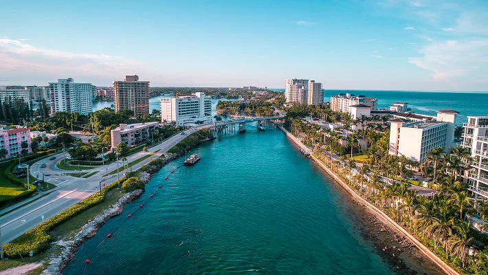 An aerial perspective looking down a vibrant blue coastal canal lined with palm trees and modern white apartment buildings, with a small bridge spanning the water in the distance.