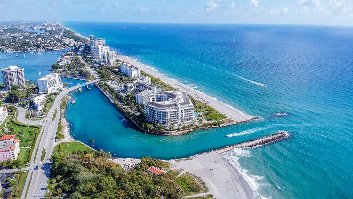 Aerial view of a coastal inlet featuring a wide turquoise waterway that connects a calm bay to the open ocean, bordered by sandy beaches and high-rise tropical resorts under a bright sky.