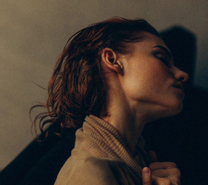 A moody, profile-view portrait of a woman with damp, reddish-brown hair. The lighting emphasizes her jawline and ear as she looks upward, wearing a textured tan turtleneck.