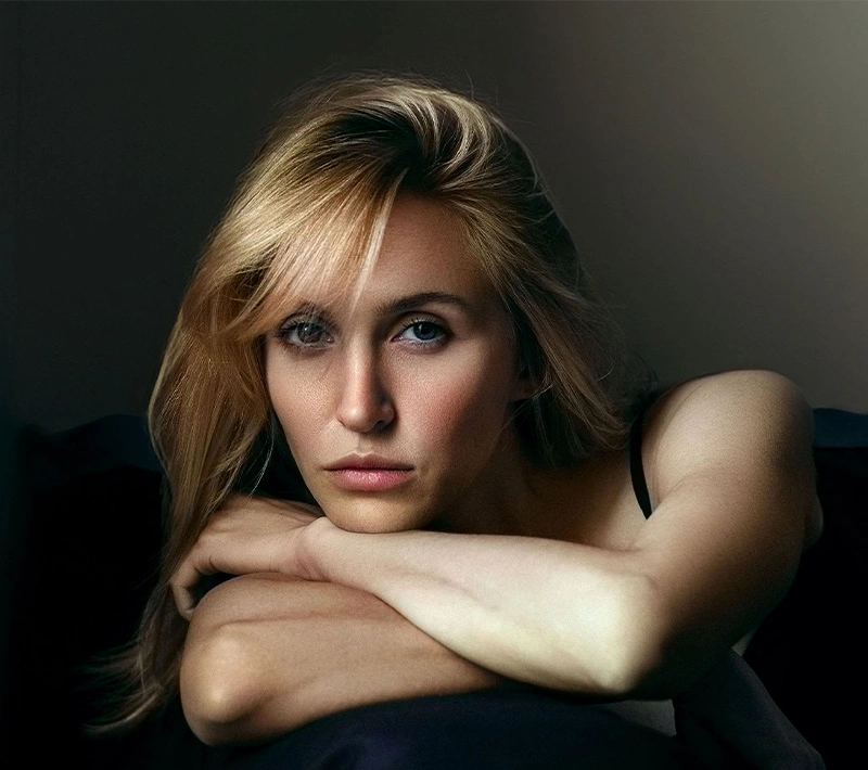 A close-up, moody portrait of a blonde woman resting her chin on her crossed arms. She has a serious, contemplative expression with soft lighting highlighting her facial structure.