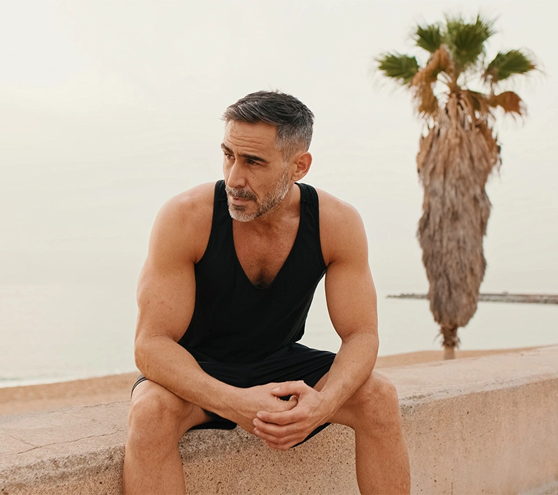 A fit, middle-aged man with salt-and-pepper hair and a beard sits on a stone wall overlooking a beach, looking thoughtfully to the side. - Align peptide blends in Los Angeles, CA