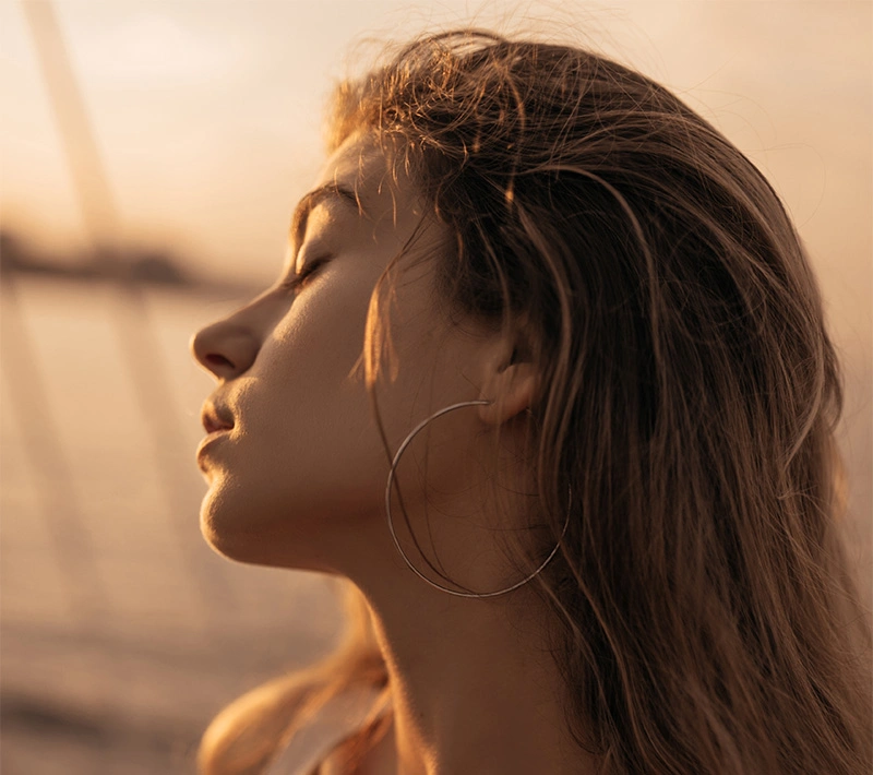 A close-up profile of a woman with large hoop earrings and sun-kissed hair, silhouetted against a golden sunset by the water with her eyes closed. - B12 in Los Angeles, CA