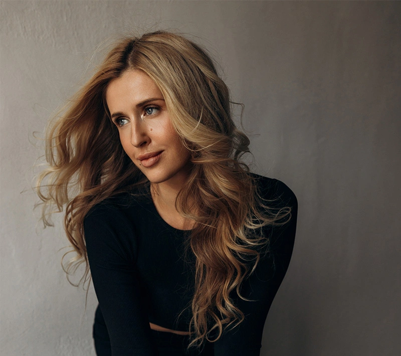 A studio portrait of a woman with long, voluminous blonde waves wearing a black long-sleeved top, looking off-camera against a textured grey backdrop. - Biostimulators in Los Angeles, CA