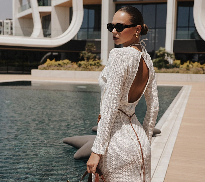 A woman in a white knit backless dress and dark sunglasses stands with her back to the camera, looking over her shoulder toward a modern poolside setting. - BBL in Los Angeles, CA