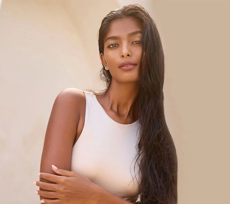 A portrait of a woman with long, dark, wavy hair and olive skin wearing a high-neck white top, posing against a neutral, minimalist background in soft natural light. - Breast implant exchange in Los Angeles, CA