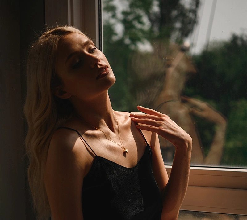 A blonde woman in a black silk camisole stands by a window in soft, moody lighting, touching her collarbone while looking down with a reflective expression. - Breast implant removal in Los Angeles, CA