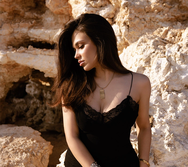 A profile view of a woman with long dark hair posing against a rugged, light-colored rock formation, wearing a black lace-trimmed camisole and a gold rectangular pendant. - Breast lift and augmentation in Los Angeles, CA