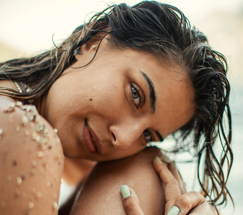 A woman with dark, flowing hair and a gold bracelet reclining on a textured light-colored blanket, with soft, dappled sunlight and shadows creating a warm, relaxed mood. - COQ10 in Los Angeles, CA