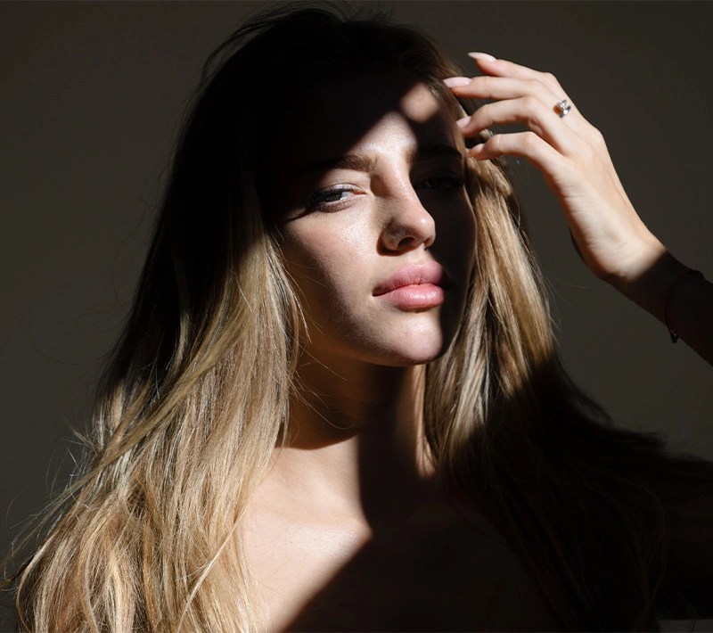 A woman with natural, glowing skin looking over her shoulder toward the camera, seated on a rocky coast with the sparkling ocean and bright sunlight reflecting in the background. - Microneedling in Los Angeles, CA