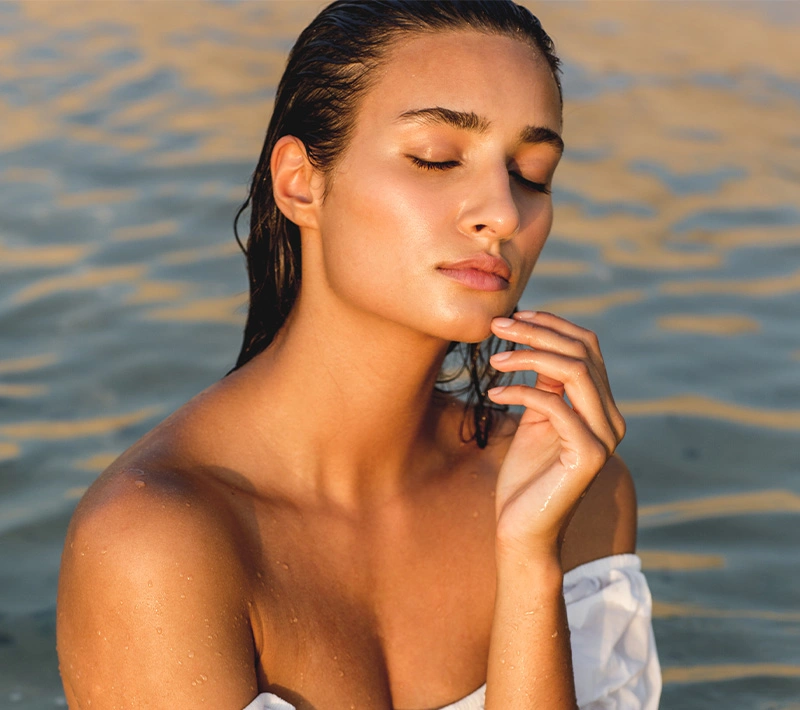 A woman with natural, glowing skin looking over her shoulder toward the camera, seated on a rocky coast with the sparkling ocean in the background. - Intro TRL in Los Angeles, CA