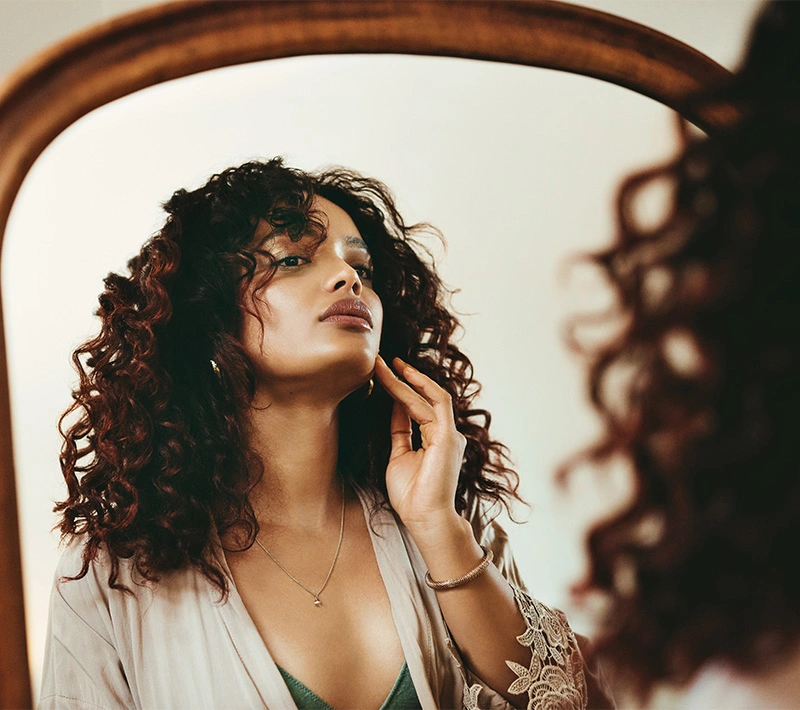 A woman with voluminous curly dark hair looking at her reflection in a vintage wooden mirror, gently touching her jawline and neck area.