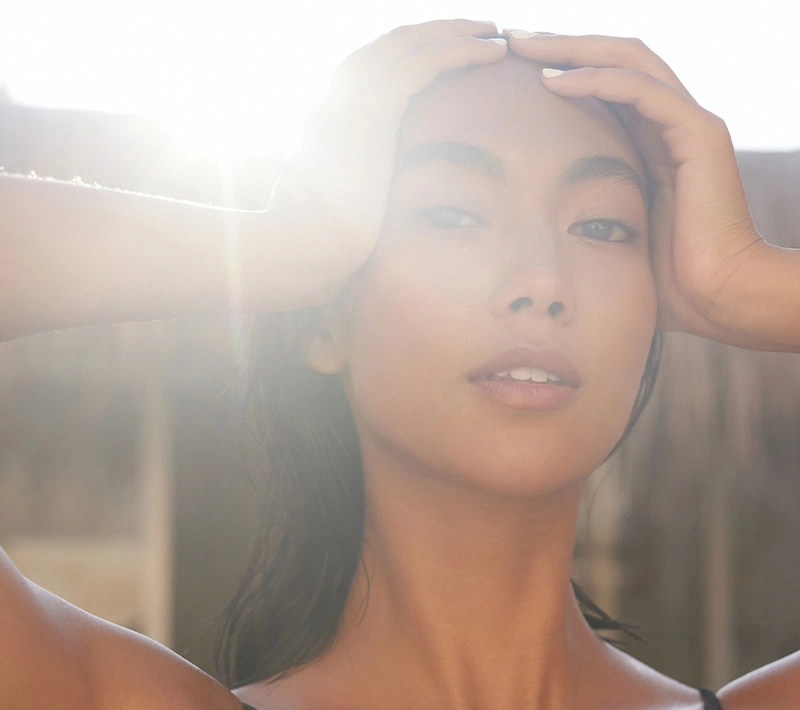 A serene portrait of a woman with radiant skin and her eyes closed, featuring the delicate shadows of a palm frond cast across her face in bright, natural light. - Pigmentation in Los Angeles, CA