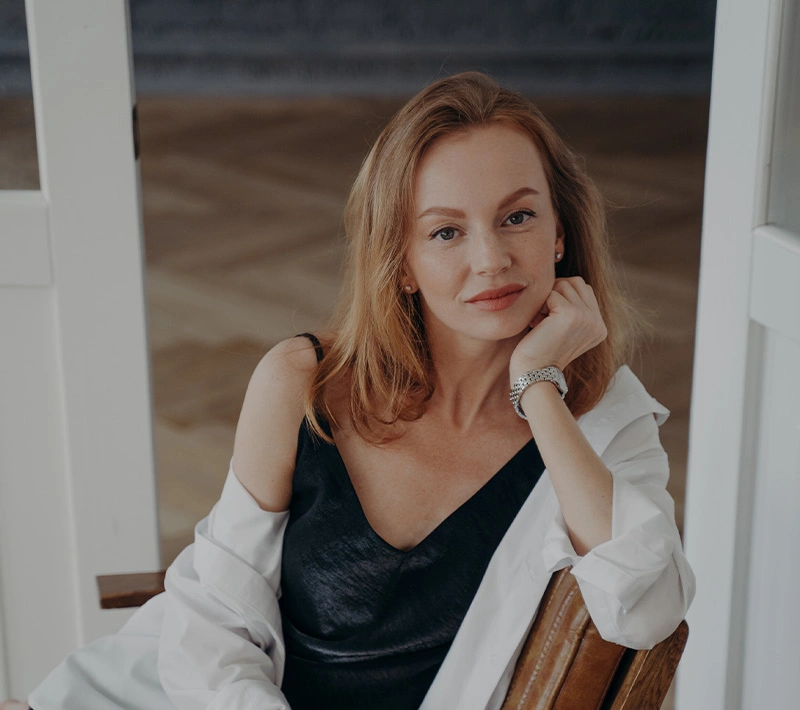 A portrait of a woman with reddish-blonde hair and freckles, wearing a black slip dress and a white button-down shirt, sitting in a bright, airy room with her chin resting on her hand.