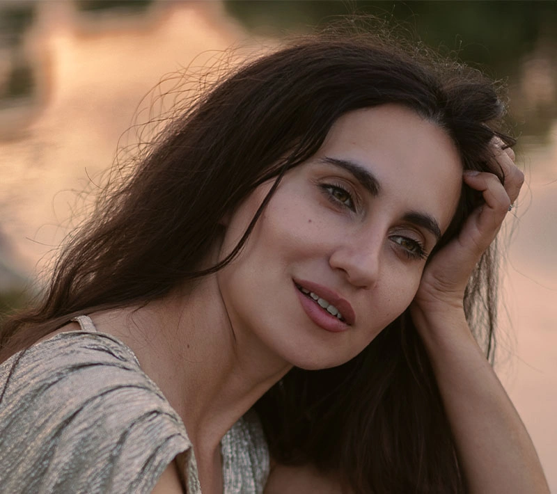A warm, close-up portrait of a woman with dark hair smiling softly and resting her hand on her head, captured during the "golden hour" with soft sunlight in the background.